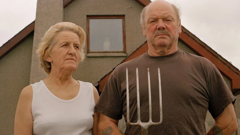 An older man and woman stand in front of their farmhouse on a gray day. The man is holding a pitchfork prongs up and staring at the camera. The woman is staring off in the distance.