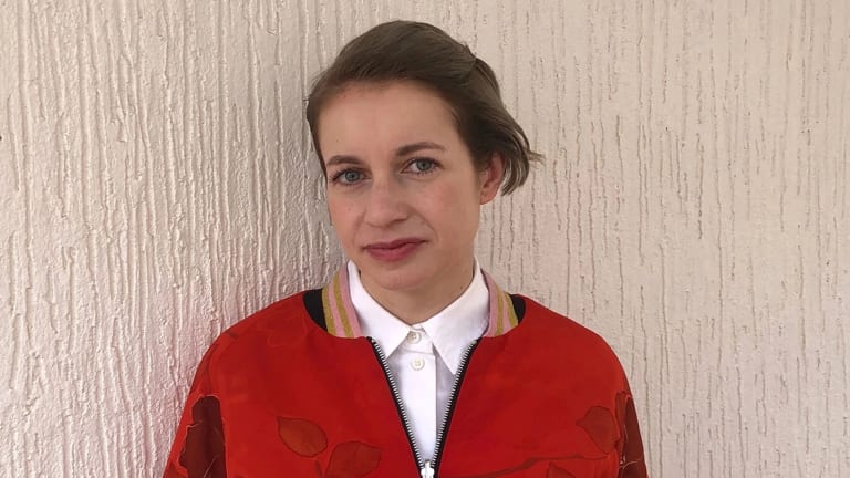 Image of Anca Rujoiu, a woman in a red jacket in front of a beige wall looking at the camera 