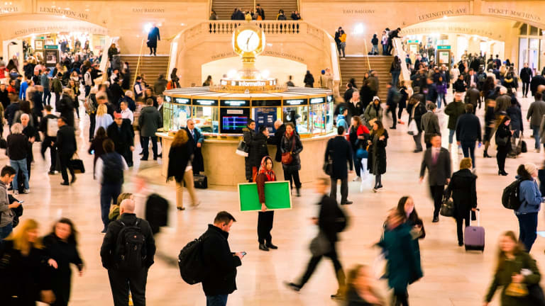 A photo of Grand Central's Main Concourse, with many people bustling around an information booth that's topped with a clock. One person in the middle of the shot is still and in focus, looking at the viewer and holding a bright green canvas under their arm.