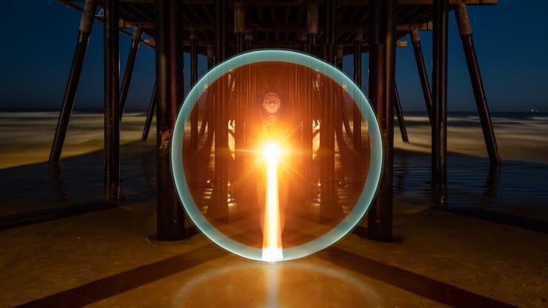 A color long exposure photograph of Russell Brown under a pier at night. He is pointing a light straight at the camera which is creating a flare in the shape of a perfect circle, framing his whole body.