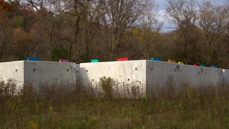 A photograph of a cement wall in a rural forest landscape. On top of the wall are multi-colored pieces of wood that make a repeating pattern.