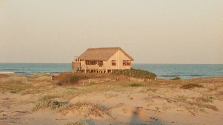 A tan house sits on a sand dune with the ocean visible in the background.
