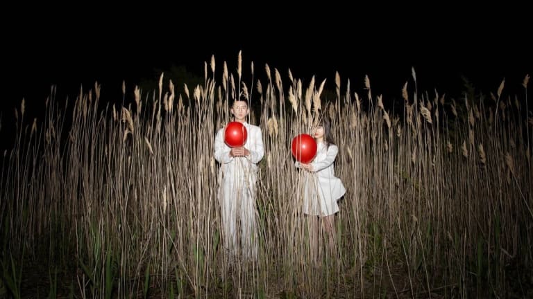 A man and woman wearing all white are each holding a red balloon, standing in tall reeds at night.