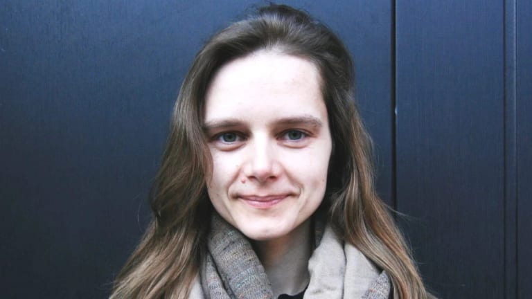 A woman with long brown hair and a gray infinity scarf on looks at the camera (Inga Lāce)