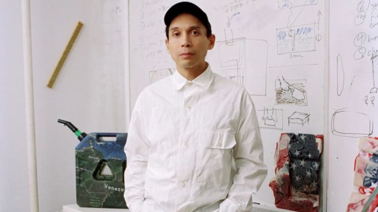 A man standing in a room with a whiteboard behind him and a green chair to his left. He's wearing a white shirt and black pants.