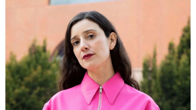 Image of a white woman in a pink zip up jacket with red lipstick and long brown hair looking at the camera with her head tilted 