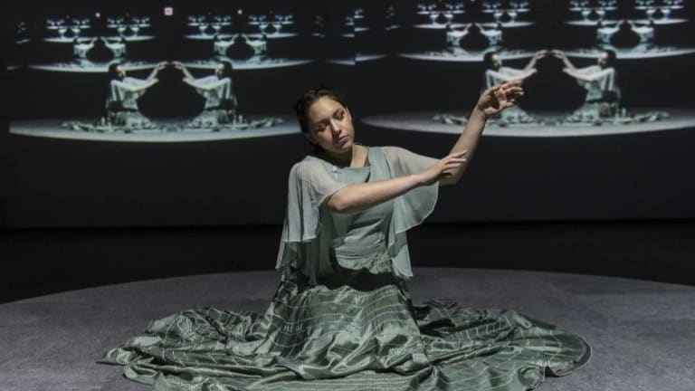 Still from performance, artist named Kite is seated and has her arms raised, with projections of her doubled on either side of the wall behind her.