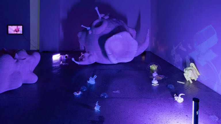 Installation by Kylie Rah. The installation is set in a large room. A large foam sculpture sits in the center of the room and is in the form of a sphere with protruding attachments. There are other small sculptures spread out across the flor. A single dark blue-violet light sits in the corner shining on the objects. The overhead lights are off.