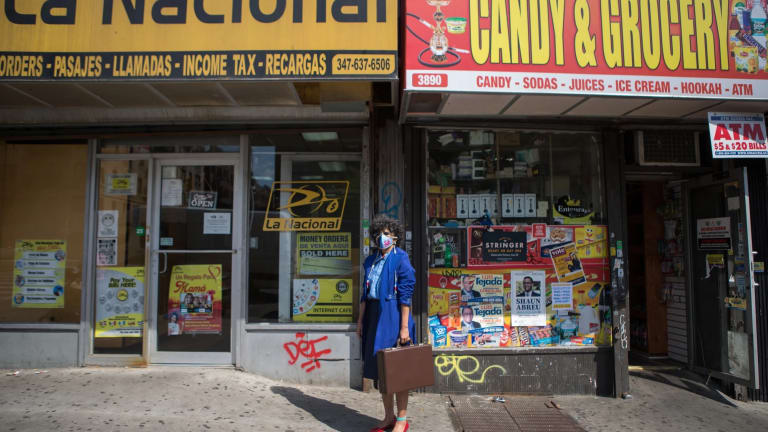 Image of a woman wearing a face mask, sunglasses, a blue coat, red shoes, and holding a briefcase standing in front of two stores with awnings that read "La Nacional" and "Candy & Grocery"