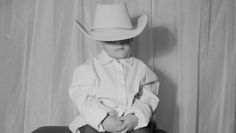 Black and white photo of a child wearing a cowboy hat that is too big for them, obscuring their face.