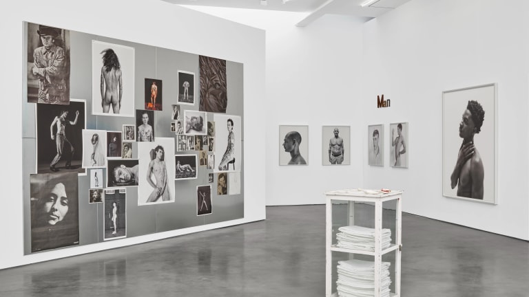 White gallery room with photographs hung in a salon style on left wall; framed portraits on right wall. Sculpture in the center of the room