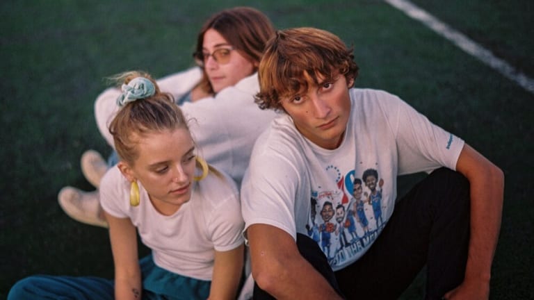 Three teens wearing white T-shirts sit on a football field in a cluster with moody looks
