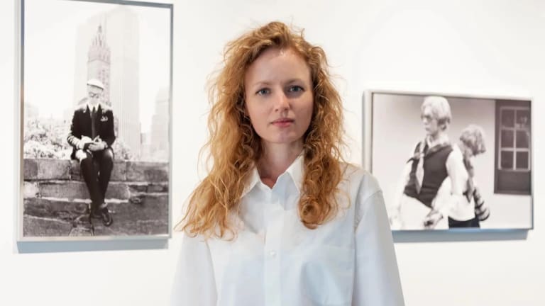 A woman with red hair stands in a gallery, behind her is a wall with two black and white photos on it 