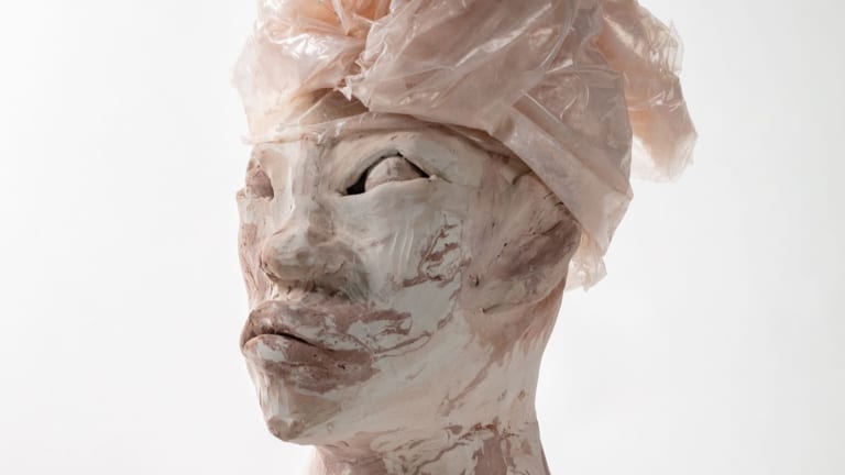 A sculpture by Symia McKellar. The sculpture is a human head with neck. The sculpture has a surface of marbled pink and white with a textile wrapped head piece.