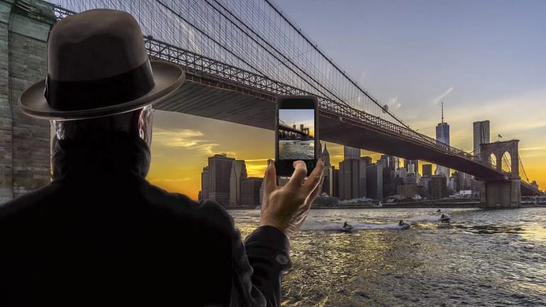 A man, wearing a brown fedora and a black coat, whose back is turned toward us, is looking across the Brooklyn Bridge at the Lower Manhattan skyline against the sunset. His right hand is raised as he is taking a photo of the skyline on his cell phone.