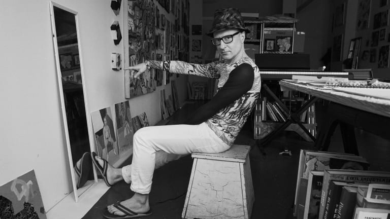 Black and white photo portrait of the artist Wayne Koestenbaum pointing at a painting in his studio.