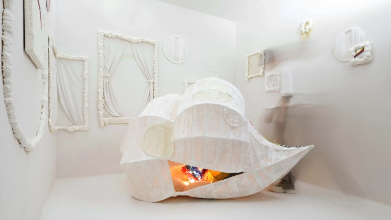An installation in a small white room of a white cloth pod, warm light glowing within, and empty frames of white fabric on the walls.