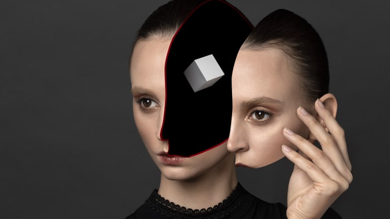 A studio headshot portrait of a woman against a gray background, wearing a black blouse. The left portion of her face from forehead to mouth has been removed, leaving a black negative void. Suspended in the void is a small, white cube. The woman’s right hand is held up next to her head, and holding the removed portion of her face like a mask.