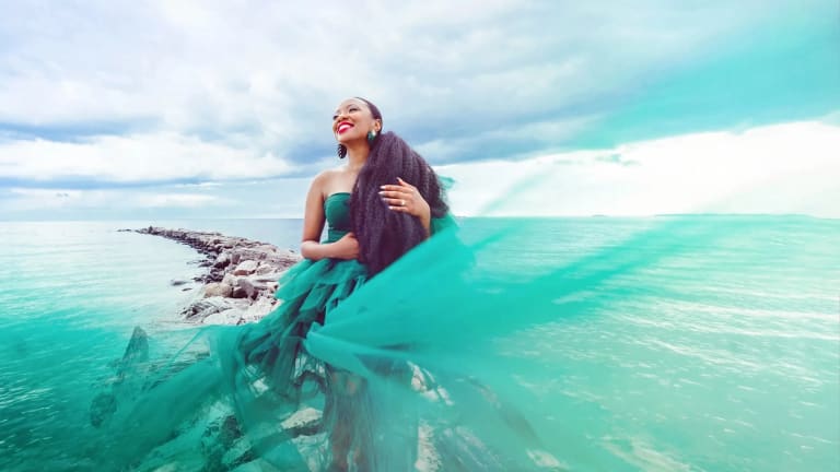 A woman in a teal dress stands smiling on a rock jetty, surrounded by the water.