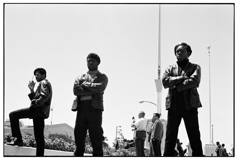 A B&W photograph of three members of the Black Panther Party in Black leather jackets Black berets standing guard outside with a flagpole in the background.