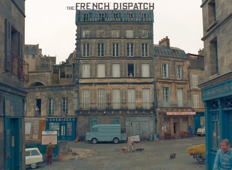 A film still showing a weather-worn building in a picturesque French town. On the roof of the building is a sign reading "The French Dispatch."