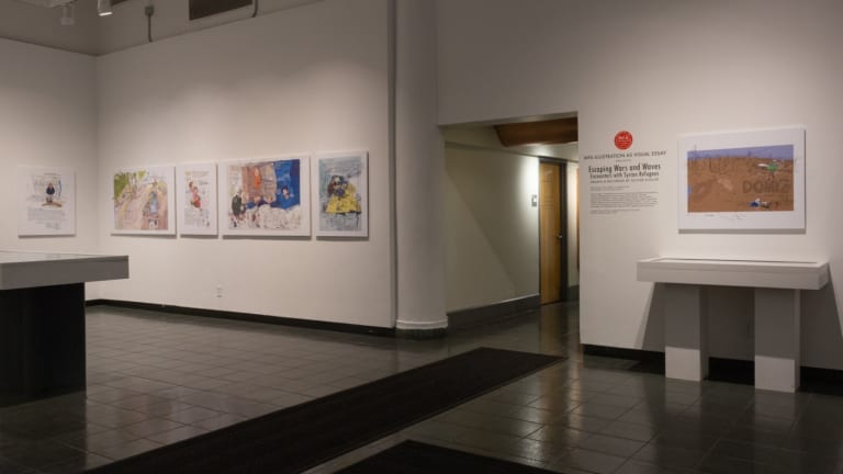 Photo of gallery immediately upon entering, with illustrated panels hung on walls and two vitrines in the space.