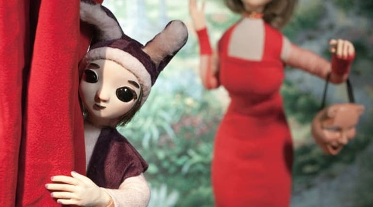 Doll wearing a hat with ears peeking around a red curtain. Doll in background wearing a red dress holding a mask.