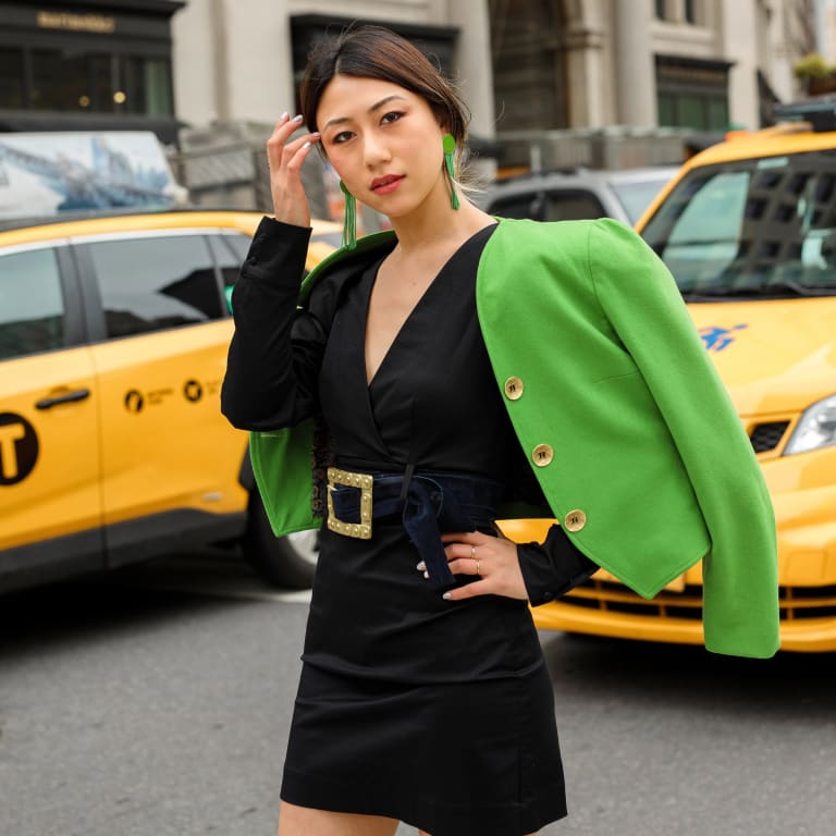 A woman in a black dress and green jacket stands confidently in front of parked taxis on a city street.