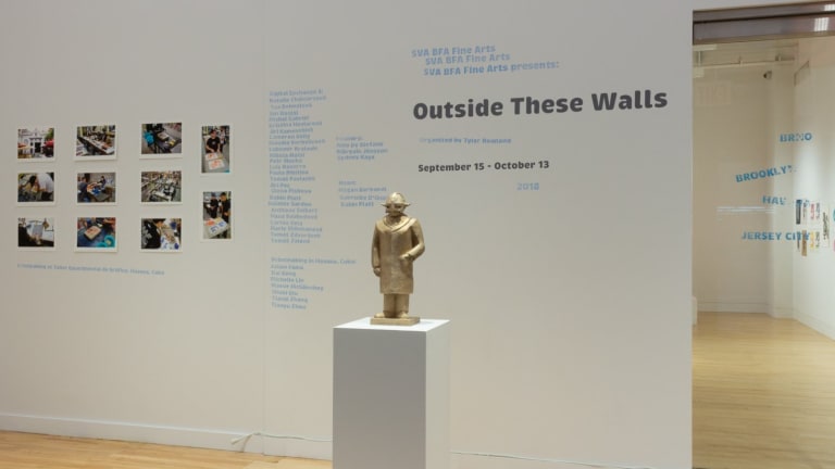 Photograph of external gallery wall with "Outside These Walls" exhibition title, artist names, and printed photographs on the wall. In the center of the room, in front of the wall, is a bronze sculpture of a Viking.