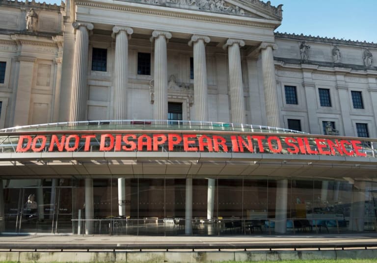 A fence weaving stating "DO NOT DISAPPEAR INTO SILENCE" is currently on view on the facade of the Brooklyn Museum as a part of the exhibition "Something to Say: Brooklyn Hi-Art! Machine, Deborah Kass, Kameelah Janan Rasheed, and Hank Willis Thomas".