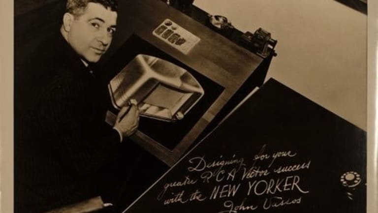 An old, black and white signed photograph of a white man sitting at a desk working on a drawing with a pen in his hand.