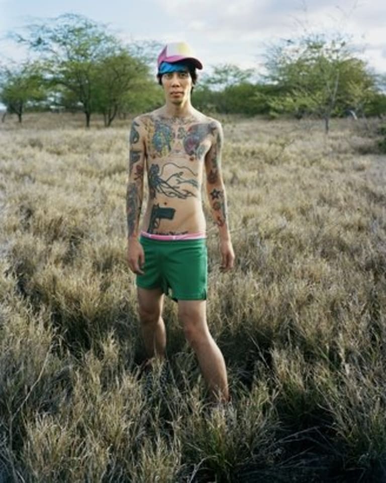 A man wearing a cap with tattoos all over his upper body in green shorts.