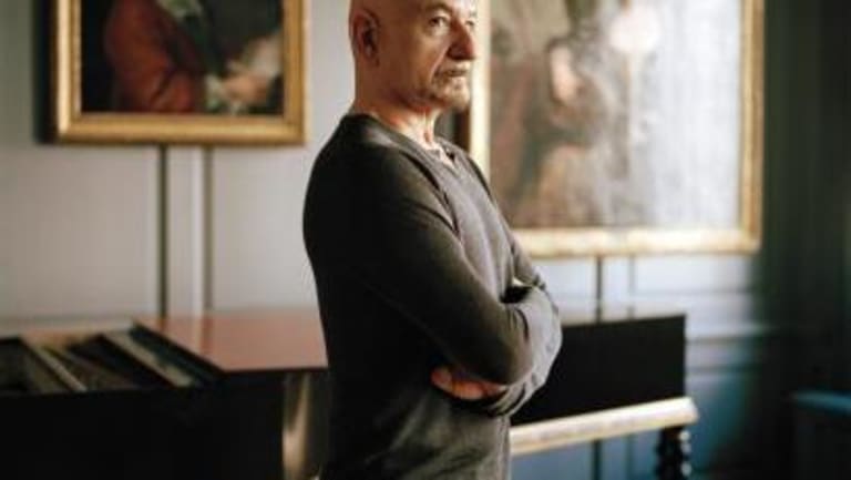 bald man with facial hair wearing long sleeve shirt and jeans in front of gold framed paintings of two people