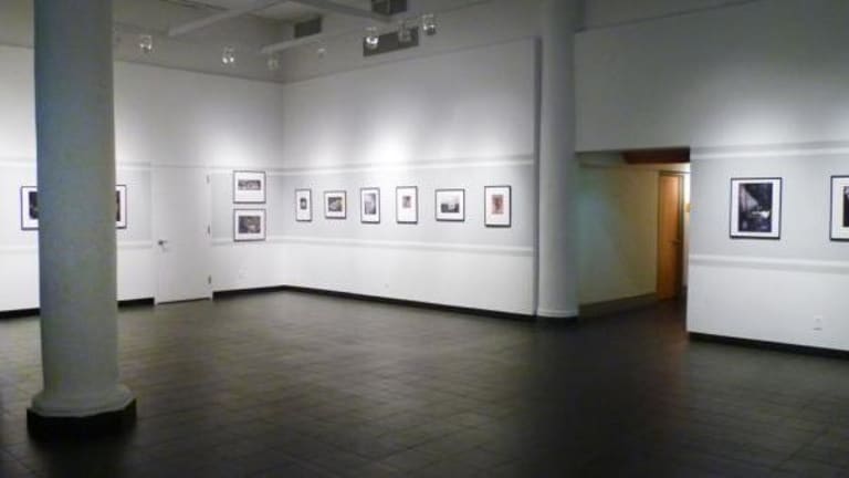 A semi-brightly lit room with photographs hanging on the white walls. There is a hallway leading elsewhere.