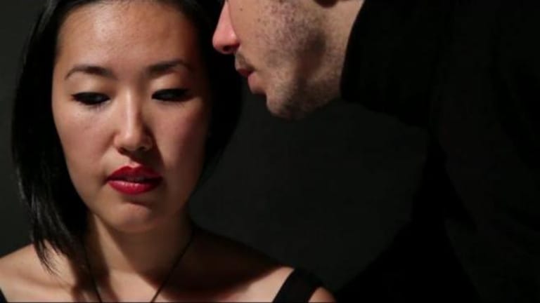 A man is whispering in the ear of a scared-looking woman with black hair and full red lips.