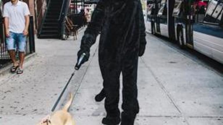 A person in a black cat costume walking a dog in a city.