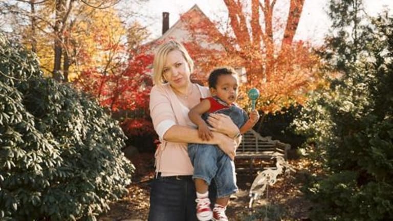 A white woman holding a black child outside in autumn. In the background, a house, a bench, and various trees.