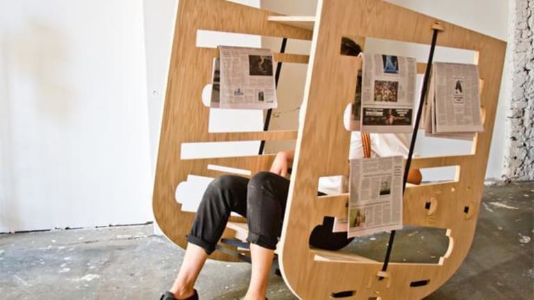 A person sits on a unique chair.  The chair has slots with news papers hanging on the slots.