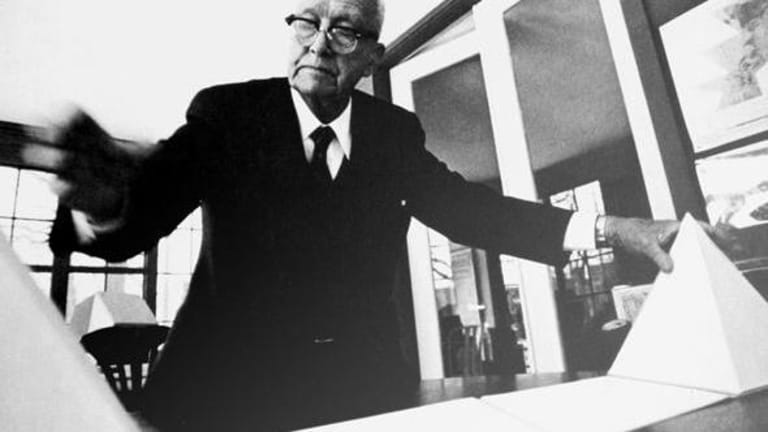 An older gentleman in a dark suit stands over a table top holding onto a white pyramid.