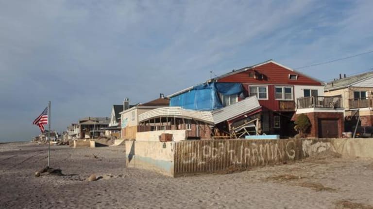 There is a sign on a brick wall that says global warming by the beach, on the sand a flag stand to the left that is an Americ flag. Several homes look that they may have been damaged in the storm one of the houses has tarps covering its windows.