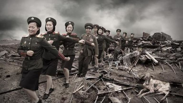Several women standing in the midst of a bunch of rubble, in military uniforms pointing with one arm out and one arm to the chest are marching and smiling, well the sky is gray black and white. With several male soldiers marching in between the groups of women.
