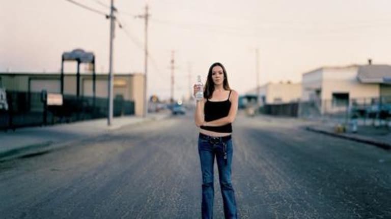 A young woman stands in the middle of a street with a bottle of alcohol in her hand.