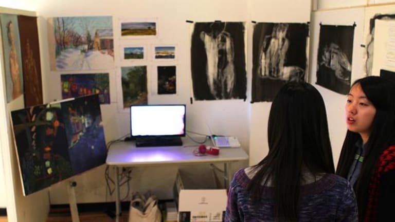 A room that shows paintings on the wall with a computer on a table. There are two Asian ladies looking.