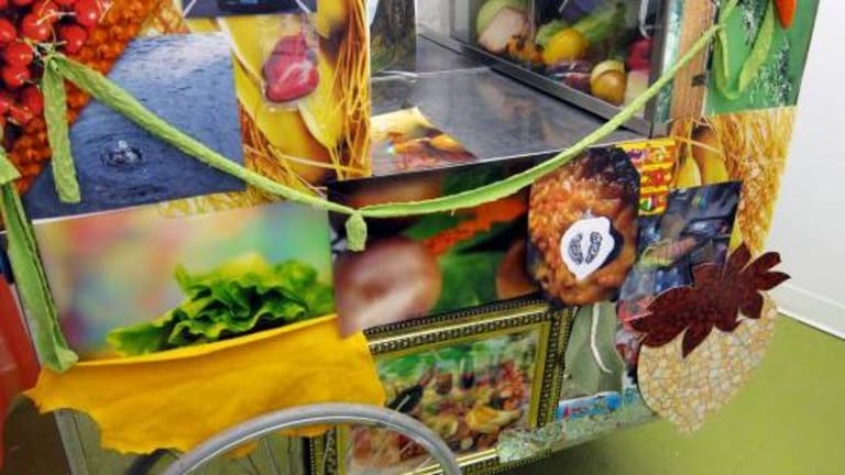 A fruit and vegetable selling cart. Fresh fruits and vegetables can be seen in the cart as well as pictures of vegetables and fruit used to decorate the cart.