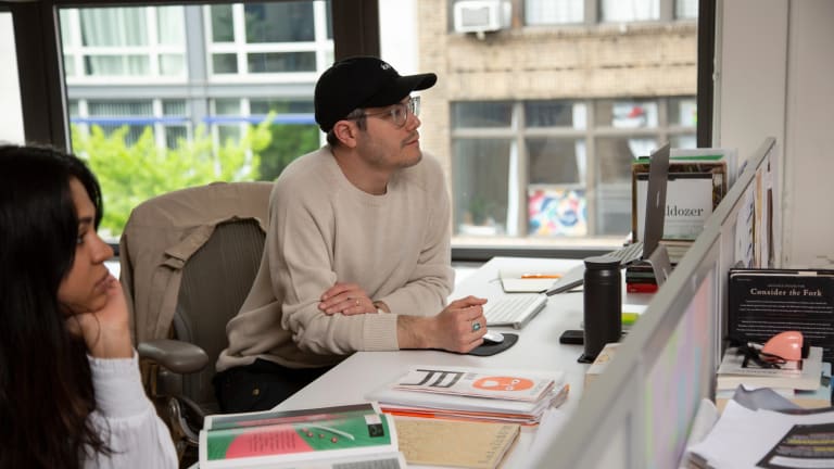 A man in a baseball cap sits at a desk in a bright room next to a woman with long brown hair surrounded by books.