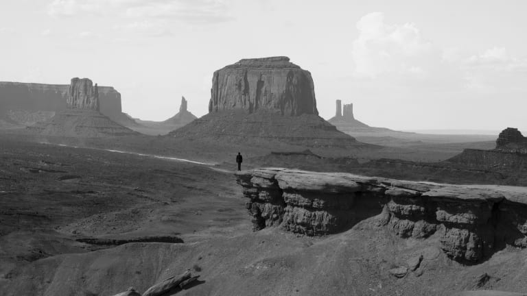 A lone silhouette stands on a cliff against a landscape of desert and mesas from the American West.