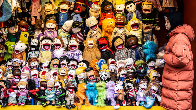 A young black girl in red winter coat walks by a street market vendor's booth, which is overflowing with large arm puppets of many various types.