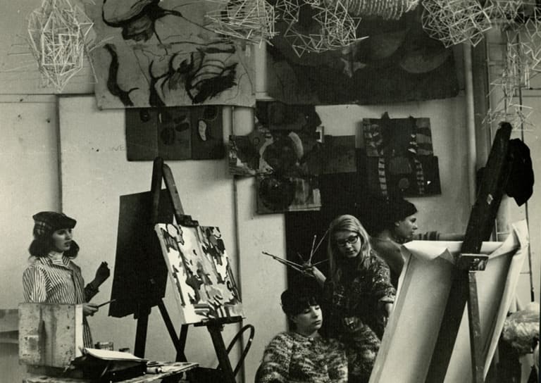 A black-and-white archival photograph of four women painting in a studio, where several paintings are hanging on the wall and sculptures hanging from the ceiling.