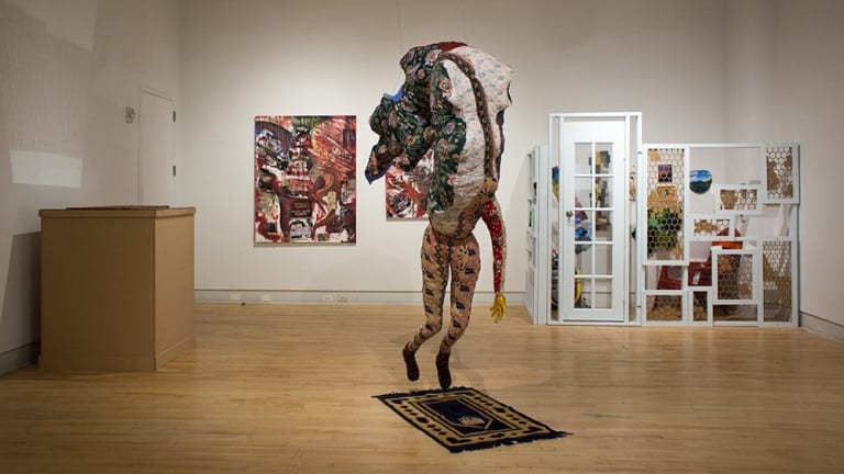 In an art gallery,  piece of art is in the middle of the room.  It is made of different patterned material with a small rug beneath it as it is suspended in the air.