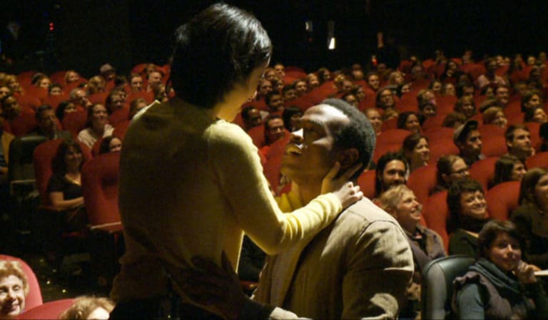 A man and a woman gazing into one another's eyes in a theater with an audience behind them